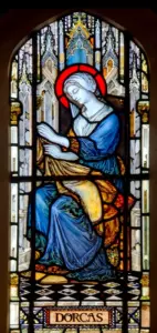Stained glass window depicting Dorcas, a haloed woman in blue and gold robes, seated and sewing a garment, with Gothic church architecture in the background.